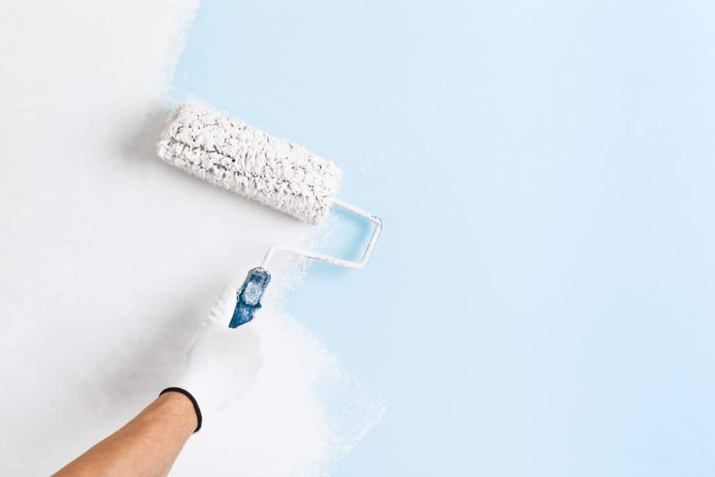 Peindre un mur : Couvrir le bleu avec du blanc | Rénovation Main gantée appliquant de la peinture blanche avec un rouleau sur un mur bleu clair. Rénovation.