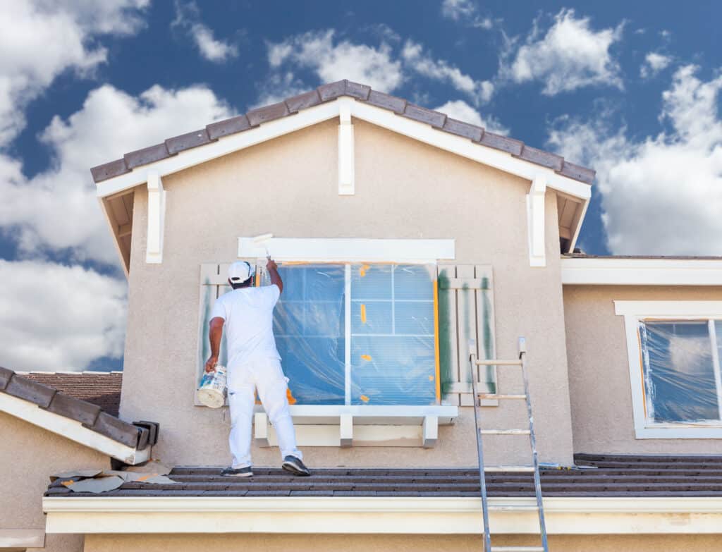 Peinture extérieure maison : ravalement façade et fenêtres Peintre en blanc sur un toit de tuiles, appliquant de la peinture blanche sur les boiseries d'une fenêtre protégée sur une façade beige. Rénovation de maison.