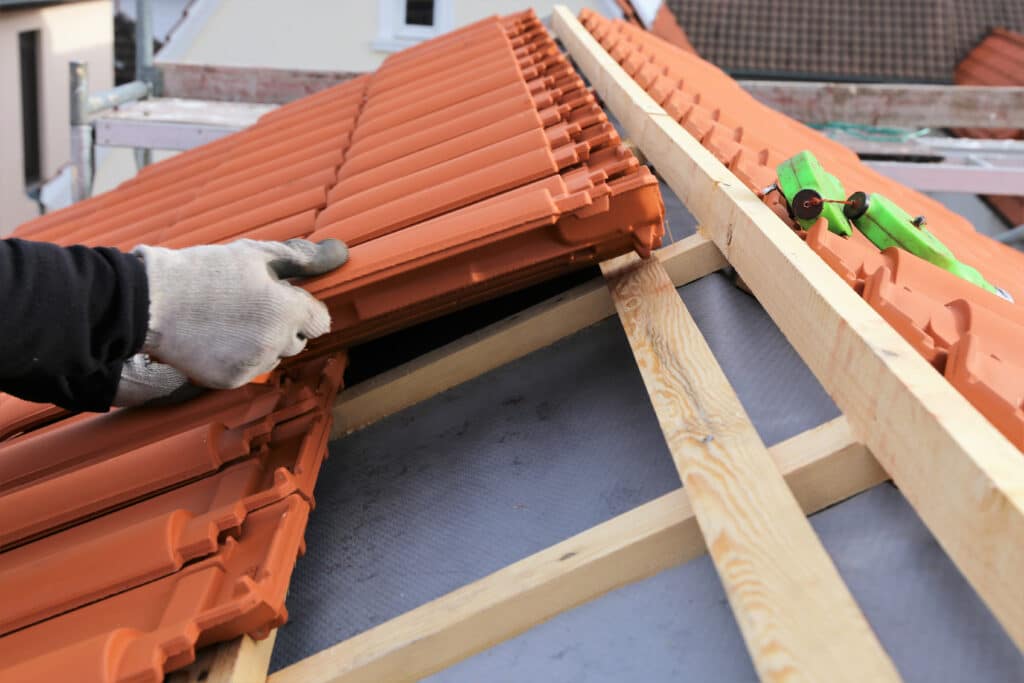 Pose de Tuiles sur Toiture : Expertise d'un Couvreur Main gantée d'un artisan posant des tuiles terre cuite sur une toiture neuve. Lattes de bois et sous-toiture gris visible. Chantier construction.