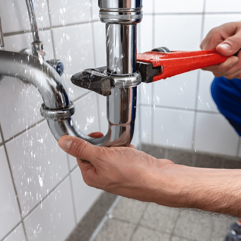 Mains manipulant une clé à molette rouge pour réparer une fuite d'eau sur un siphon chromé sous un évier, l'eau éclaboussant.